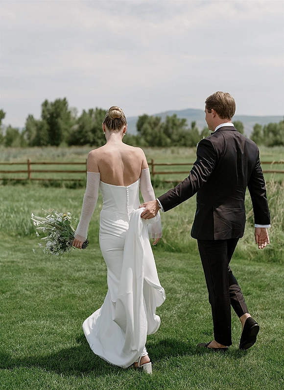 Wedding couple walking across a grassy lawn, bride holding bouquet and groom lifting her gown train near a rustic wooden fence and trees