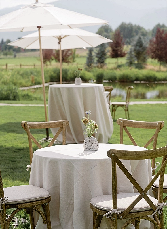 Outdoor reception seating with cocktail hour tables, white linen round table, cross-back chairs, and a wildflower bud vase by the pond