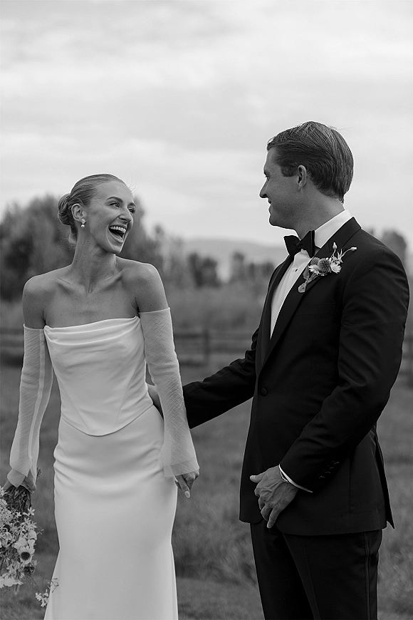 Couple portrait in a black and white wedding portrait, bride laughing in strapless gown and sheer gloves beside groom in tux by rustic fence field