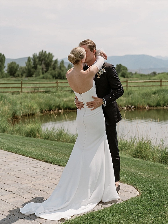 Wedding kiss portrait of bride and groom in an outdoor wedding kiss by a pond, her strapless gown train flowing with mountains behind