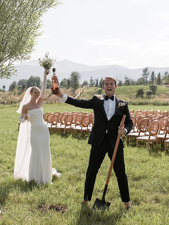 Ceremony moment as bride and groom cheering while champagne sprays from a popped bottle, bouquet and veil on a mountain lawn aisle