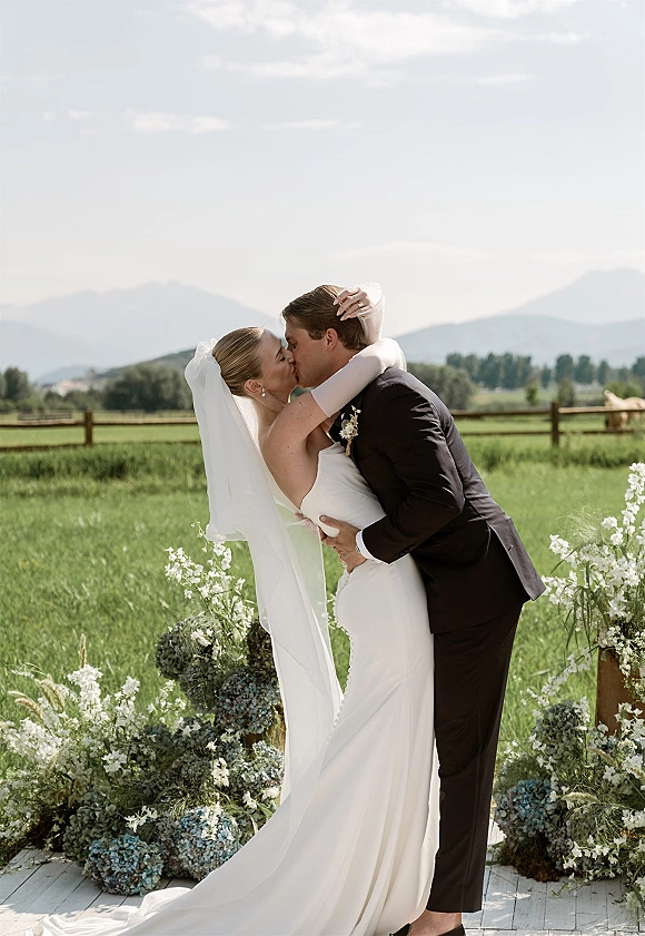 Wedding kiss portrait of bride and groom kiss, her long veil flowing as he dips her in a grassy field with mountain backdrop