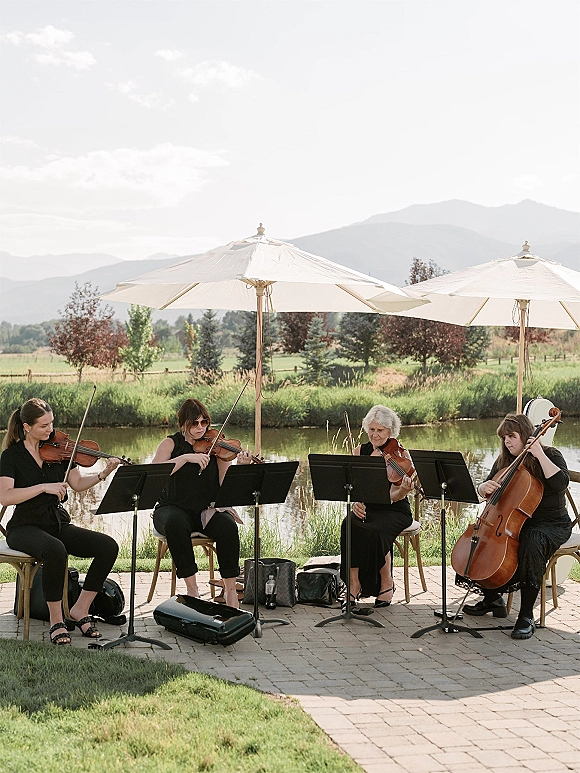 Wedding string quartet ceremony musicians playing violins and cello with sheet music stands under patio umbrellas by a pond and mountains