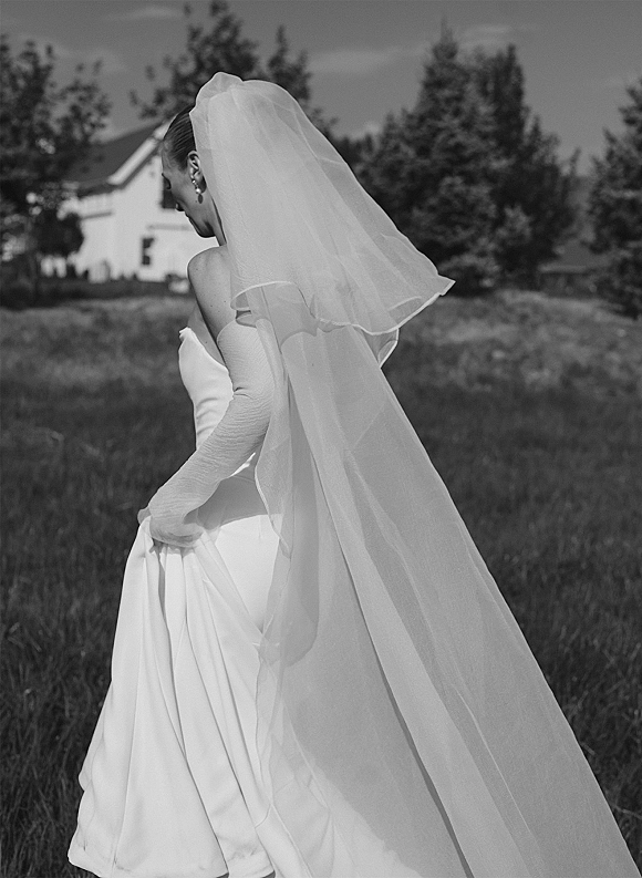 Bridal portrait of a bride from behind in a grassy field, holding her dress train with a long cathedral veil flowing behind her