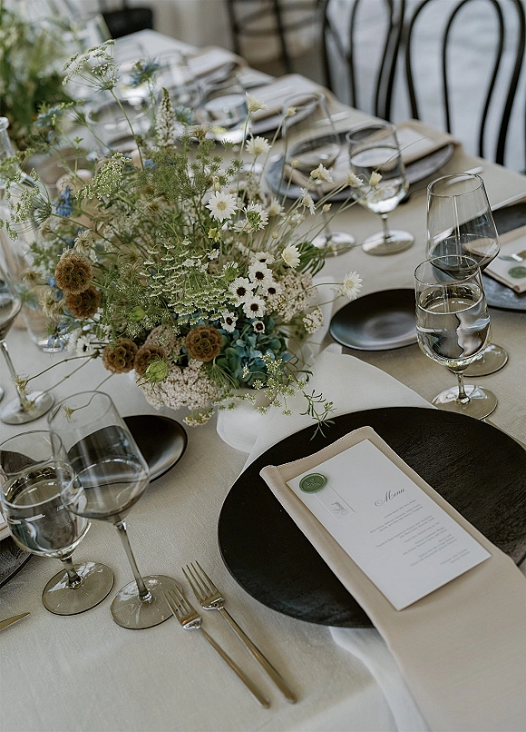 Reception tablescape with wedding table centerpiece of wildflowers on neutral linen, black charger plates, menus with wax seal, and glassware