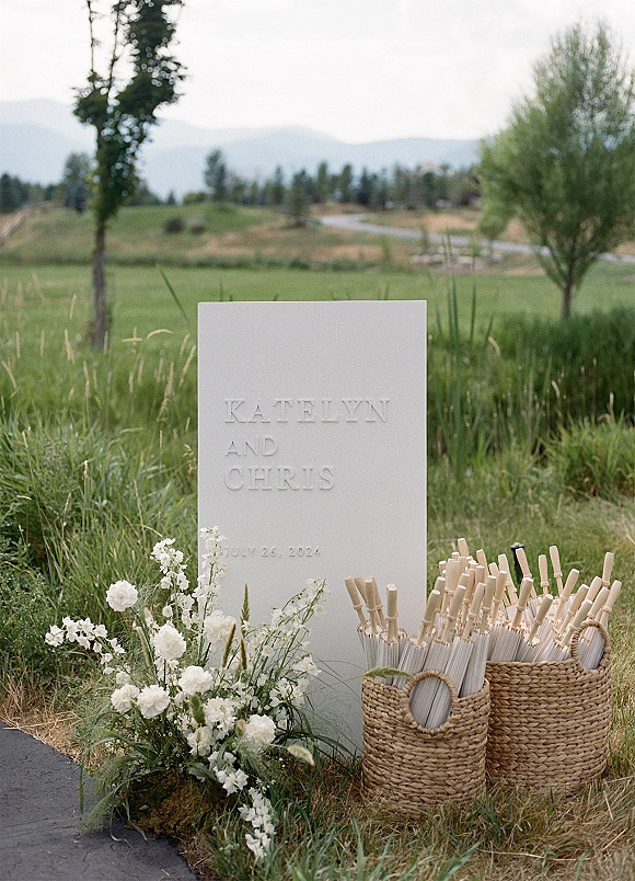 Wedding welcome sign with minimalist welcome sign styling, embossed white lettering and white flowers by woven fan baskets in a meadow