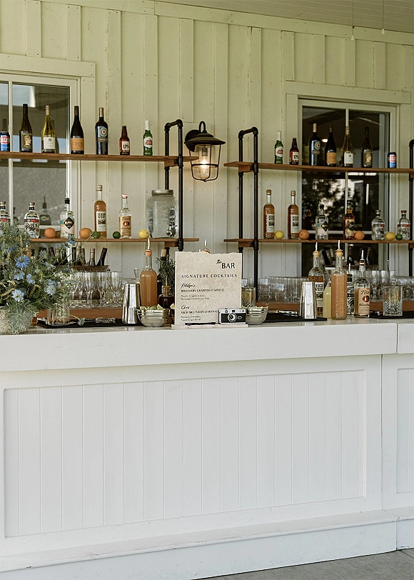 Wedding bar setup with wedding bar menu sign, liquor bottles and glassware on shelves, citrus and rosemary garnish bowls against white paneled wall