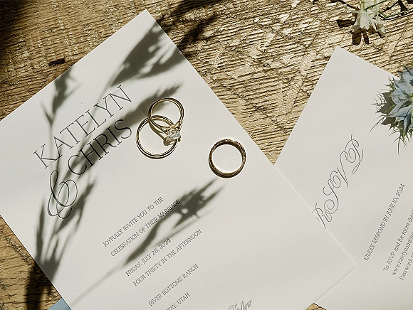 Invitation suite wedding invitation flat lay with gold wedding bands and engagement ring on printed cards, floral sprig, on wood tabletop in natural light shadows