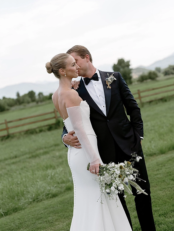 Couple portrait of bride and groom embrace as he kisses her cheek, her wildflower bouquet by her side in a green field by a fence