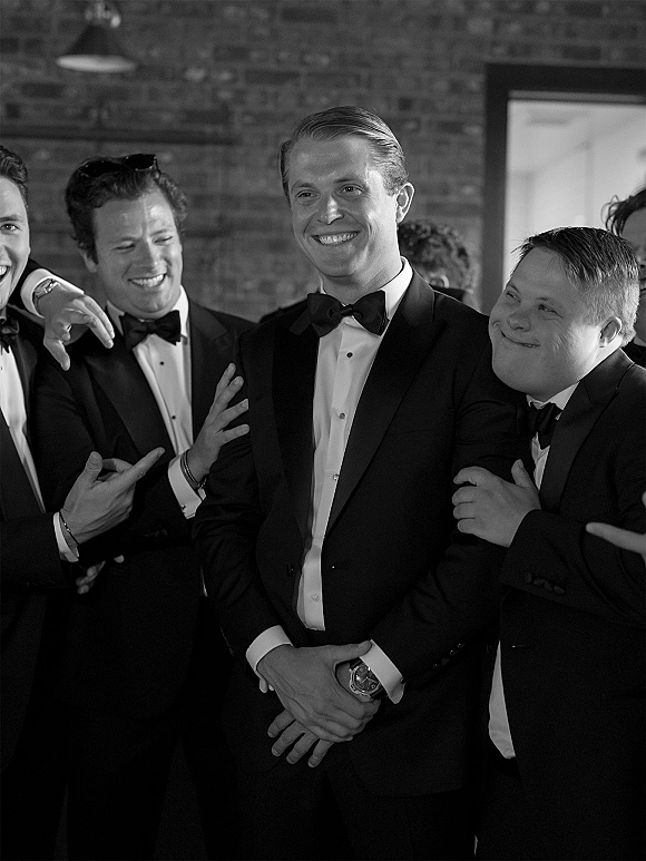 Groomsmen portrait of the groom with groomsmen in black tuxedos and bow ties, laughing by a brick wall doorway indoors