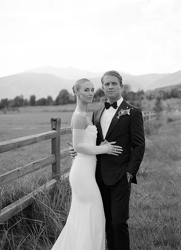 Couple portrait in a black and white wedding portrait style, bride in pearl button gown and gloves beside groom in tux by a fence in a meadow with mountains under cloudy sky