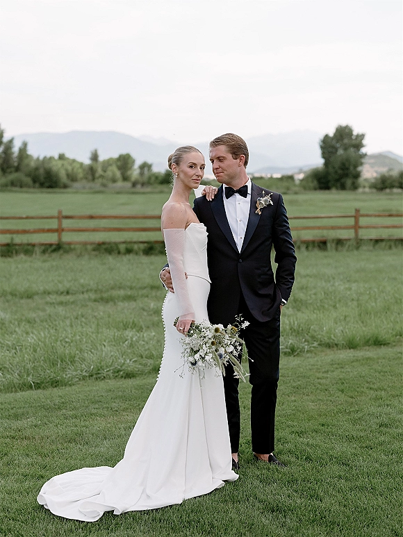 Couple portrait of bride holding bouquet beside groom in a black tuxedo, her strapless gown with long train in a mountain field under clouds