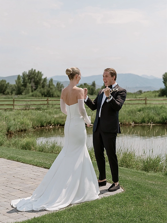 First look moment as bride in a strapless gown with long train holds hands with groom in black tuxedo by a pond with mountains behind