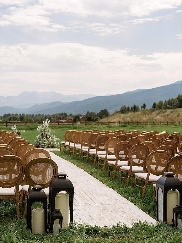 Ceremony setup with outdoor ceremony seating of wooden chairs and white cushions lining an aisle runner with black lanterns on a mountain lawn