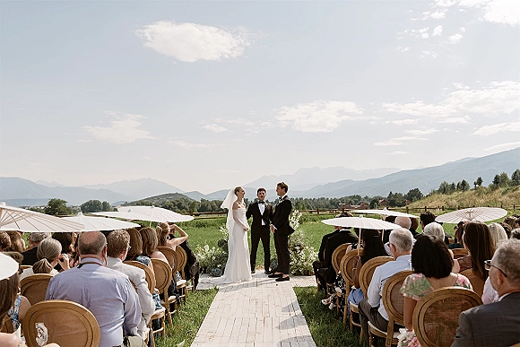 Outdoor wedding ceremony with a mountain wedding ceremony view, bride and groom at the altar with white parasols on a grassy field