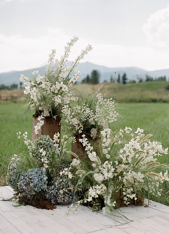 Ceremony floral arrangement of grounded ceremony florals with white wildflowers and greenery on wooden pedestals in a grassy field with mountain view
