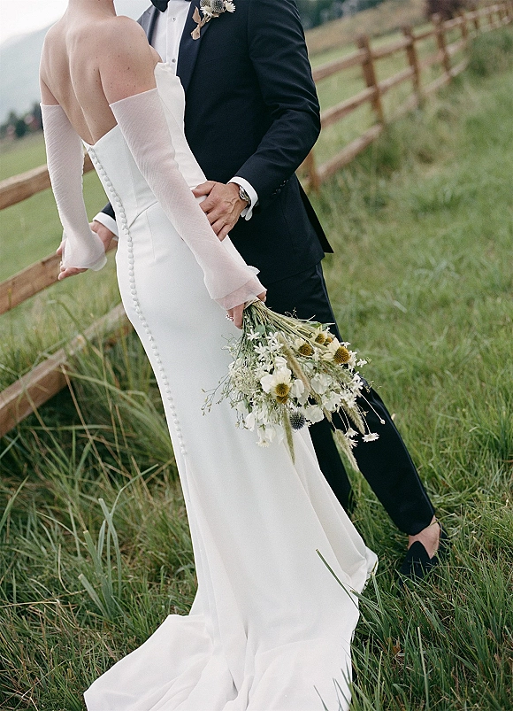 Couple portrait of bride and groom embrace, she holds a wildflower bouquet with thistle accents in a grassy field by a wooden fence