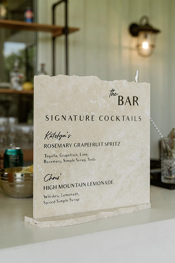 Bar menu sign with signature cocktail sign printed on stone, set on a wedding bar counter with bottles, cans, and an ice bucket behind