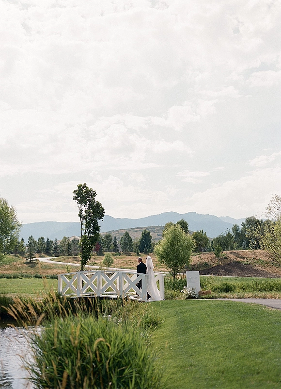 Couple portrait of bride and groom on bridge, walking away with veil train, white wooden bridge by pond and mountains under cloudy sky