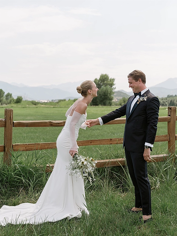 Couple portrait of bride and groom laughing, holding hands as she carries a bouquet in tall grass with mountains under a cloudy sky