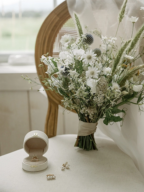 Bridal bouquet of white daisies and blue thistle with greenery, ribbon wrap, ring box and pearl earrings on a chair in window light