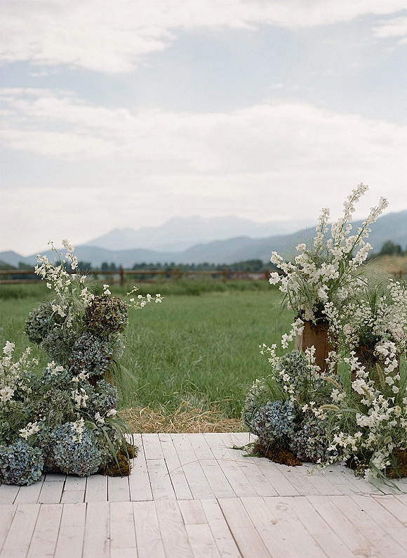 Ceremony altar decor with outdoor wedding altar florals of white blooms and hydrangeas on wood pedestals, set in a grassy field by mountains