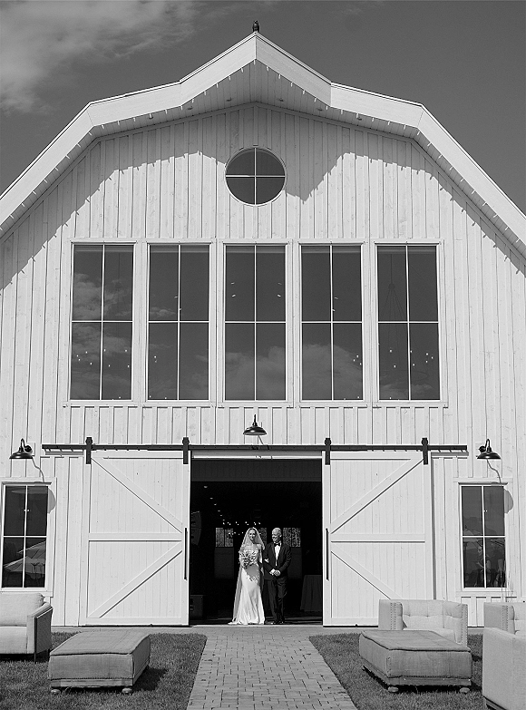 Wedding processional as bride walking down aisle with long veil and bouquet beside a tuxedoed escort at white barn doors on brick walkway