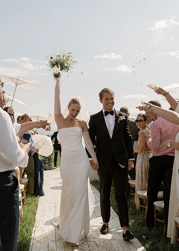 Wedding recessional as newlyweds walking down aisle hand in hand, bride raising bouquet under veil while confetti falls and guests cheer outdoors