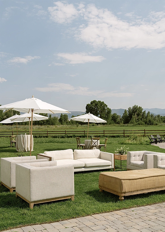 Outdoor lounge setup with wedding lounge furniture, white patio umbrellas, sofa and armchairs around an ottoman on a paver patio with mountain views