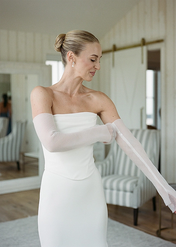 Bridal portrait of a bride getting ready in a strapless satin gown, adjusting sheer opera gloves in a bright room with white paneled walls