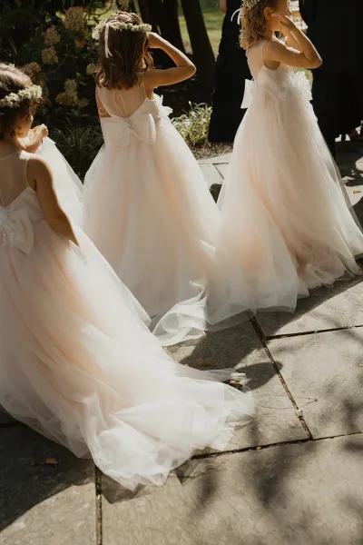 Flower girls in tulle flower girl dresses with satin bows and a floral crown, walking on a sunlit stone patio amid garden greenery