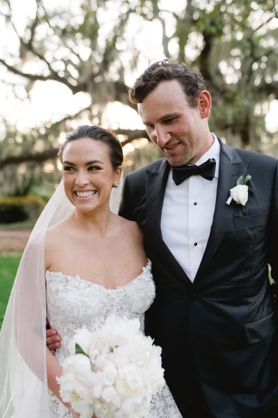 Couple portrait of bride laughing in a lace dress and veil holding a white bouquet, embracing groom in black tuxedo at sunset lawn