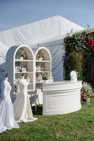 Wedding display decor with wedding welcome display shelves, framed photos, floral arrangements, and gown mannequins inside a white tent on grass