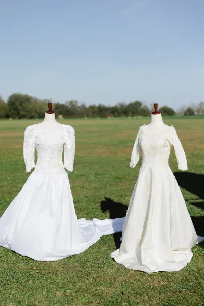 Wedding dresses on mannequins, including a lace wedding dress with long sleeves, set in a grassy field with trees under blue sky
