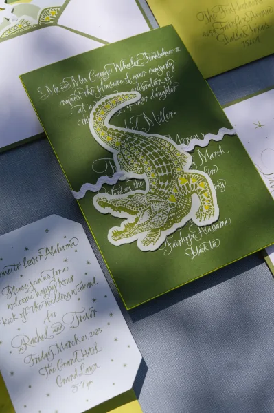 Wedding invitation suite featuring green wedding invitations with calligraphy lettering, alligator illustration, and gold ink on blue fabric backdrop