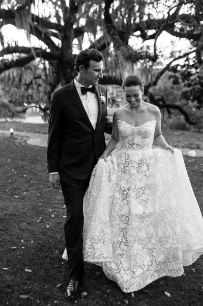 Couple portrait in a black and white wedding portrait as bride lifts her lace gown while walking with groom in tuxedo on a garden path