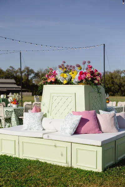 Reception lounge seating with an outdoor wedding lounge sofa, throw pillows, and floral arrangement under string lights on a grassy lawn