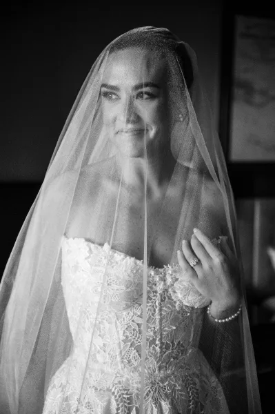 Bridal portrait of a bride under veil in window light, wearing a strapless lace dress and drop earrings against a dark backdrop