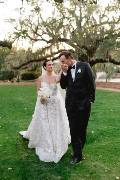 Couple portrait of groom kissing bride’s hand as they walk on a green lawn under moss-draped trees, bride in lace dress holding bouquet
