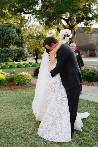 Wedding kiss as bride and groom embrace, her lace gown and long veil flowing while she holds a white bouquet on a garden lawn
