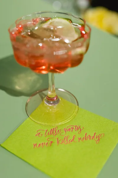 Signature wedding cocktail in a stemmed glass with ice and citrus slice garnish on a calligraphy wedding cocktail napkin atop a glass tabletop