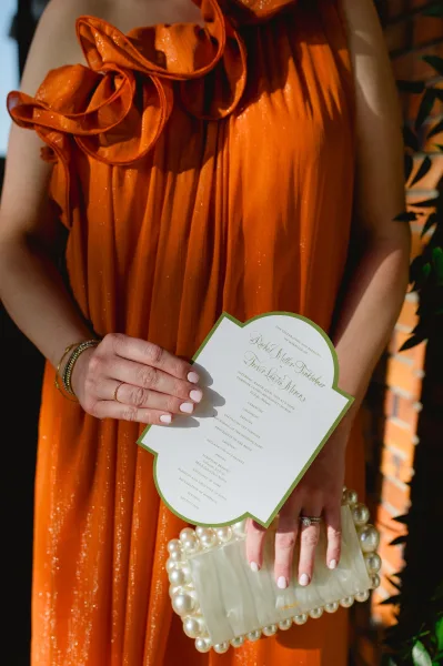 Wedding invitation and wedding ceremony program held by a bridesmaid in an orange ruffle dress, with pearl clutch and gold bracelets outdoors