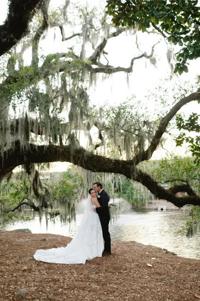 Couple portrait of bride and groom embrace under an oak tree with Spanish moss, her lace gown and veil flowing by the lake shoreline