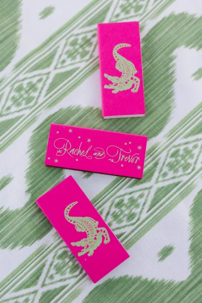 Wedding matchbooks with personalized matchbooks in hot pink, gold foil calligraphy, and alligator illustration on green patterned fabric