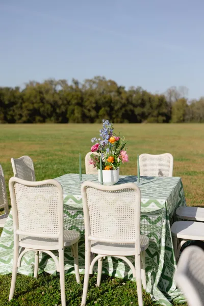Reception tablescape with an outdoor reception table, green patterned cloth, wildflower centerpiece in a white vase, and taper candles in a field under blue sky