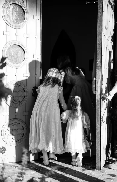 Flower girl moment as two flower girls walking in, holding hands in lace dresses with hair bows at a church doorway with carved doors