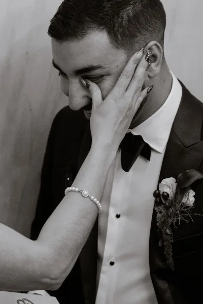 Groom portrait of a man in black tuxedo wiping tears as the bride touches his face, her pearl bracelet visible against a plain wall