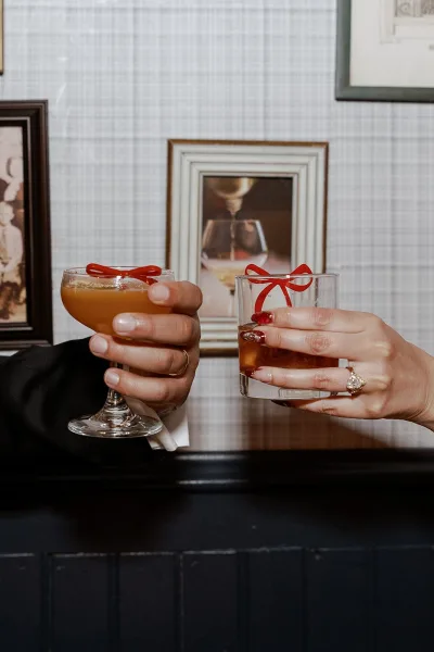 Wedding toast as couple clinks cocktail glasses with red ribbon bows, showing engagement ring and tuxedo sleeve against framed photo wall backdrop
