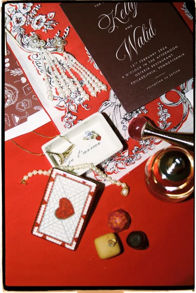 Invitation suite flat lay with calligraphy card, pearls, rings, wax seal stamp, and chocolates arranged on a red tabletop surface