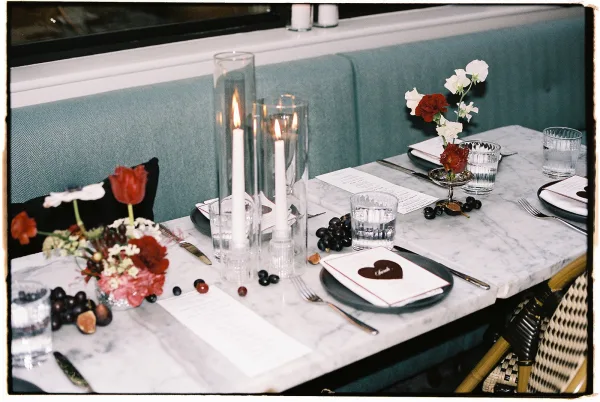 Reception tablescape on a marble wedding table with glass hurricane candles, red and white flowers, black plates, and teal banquette seating